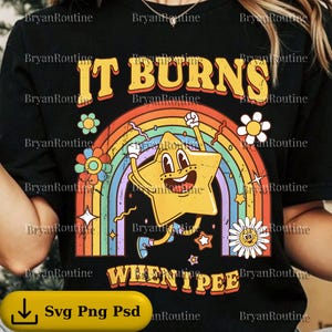 May include: Black t-shirt with a retro design featuring a yellow star character, a rainbow, and the text "IT BURNS WHEN I PEE". The design includes flowers and stars in a colorful, cartoon style.