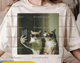 May include: Cream-colored t-shirt featuring a square image of two cats wearing sunglasses, one taking a selfie with a smartphone. The image has a green background. The text "Svg Png Psd" is at the bottom.