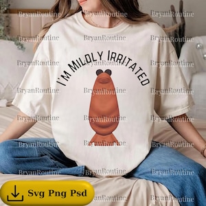 May include: Cream-colored t-shirt with the text "I'M MILDLY IRRITATED" above a cartoonish brown creature. The shirt is a casual, short-sleeved style, suitable for everyday wear. The image also includes a yellow button with the text "Svg Png Psd".