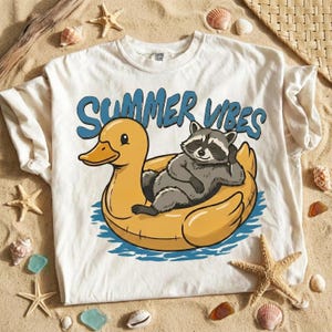 Op de afbeelding: Wit T-shirt met een afbeelding van een wasbeer die ontspant op een gele eenden-opblaasboot. De tekst "SUMMER VIBES" staat in blauw boven de wasbeer en de eend. Het shirt ligt op een zanderige ondergrond met schelpen en zeesterren.