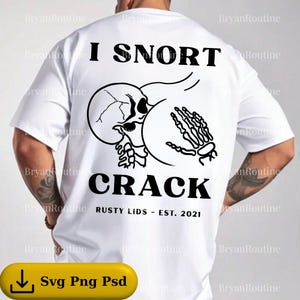 May include: White t-shirt with a black graphic design. The design features a skull, a skeleton hand, and the words "I SNORT CRACK" in bold letters. The text "RUSTY LIDS - EST. 2021" is also included.