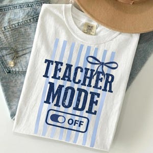 May include: White t-shirt with the text "TEACHER MODE OFF" in navy blue, accented by light blue vertical stripes and a bow graphic. The shirt is folded on top of blue jeans and a tan hat.