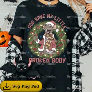 May include: Black t-shirt with a Christmas-themed graphic. The design features a cartoon rat wearing a Santa hat and coat, surrounded by a wreath and the text "GOD SAVE MY LITTLE BROKEN BODY."