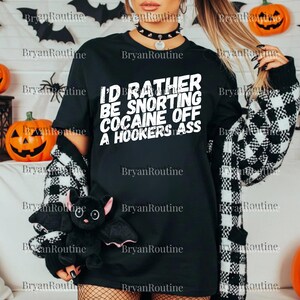 以下が含まれることがあります： 「I'D RATHER BE SNORTING COCAINE OFF A HOOKERS ASS」の白い文字が入った黒いTシャツ。黒と白のチェック柄のカーディガンを着て、黒いコウモリのぬいぐるみを持っています。背景にはハロウィーンの装飾があります。