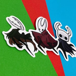 Op de afbeelding: Drie stickers met personages uit het videospel Hollow Knight. De stickers bevinden zich op een rode, groene en blauwe achtergrond. De personages zijn de Ridder, het Witte Paleis en de Zielmeester.