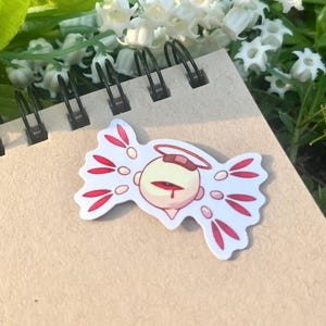 Pode incluir: Um adesivo com um personagem de desenho animado com um olho, um halo e detalhes vermelhos. O adesivo está colocado em um caderno espiral, com flores brancas ao fundo.