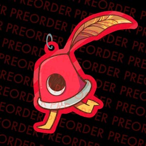 Puede incluir: Un dije rojo en forma de campana con un diseño de pluma y media luna. El dije tiene pies amarillos y un lazo plateado para sujetar. El fondo es negro con las palabras "PREORDER" repetidas.