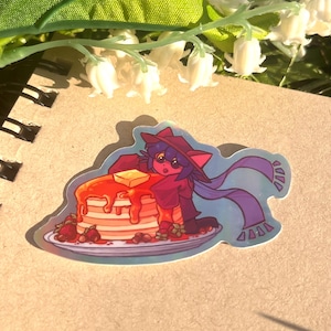 May include: A holographic sticker featuring a cartoon character with cat-like features, wearing a red hat and scarf, standing next to a stack of pancakes with syrup, butter, and strawberries on a plate.