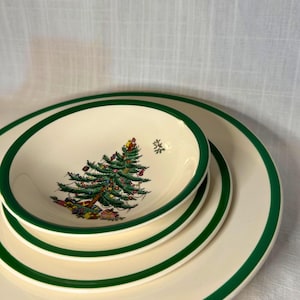 May include: A stack of off-white ceramic plates and bowls with a green border. The smallest bowl features a Christmas tree design with presents. The plates and bowls are stacked on top of each other.