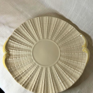 May include: A cream-colored, round serving platter with a textured, fan-like pattern radiating from the center. The platter has two yellow handles on either side. The plate is made of ceramic.
