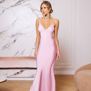 May include: A woman wearing a light pink satin maxi dress with a V-neckline and spaghetti straps. The dress has a fitted bodice and a flowing skirt.
