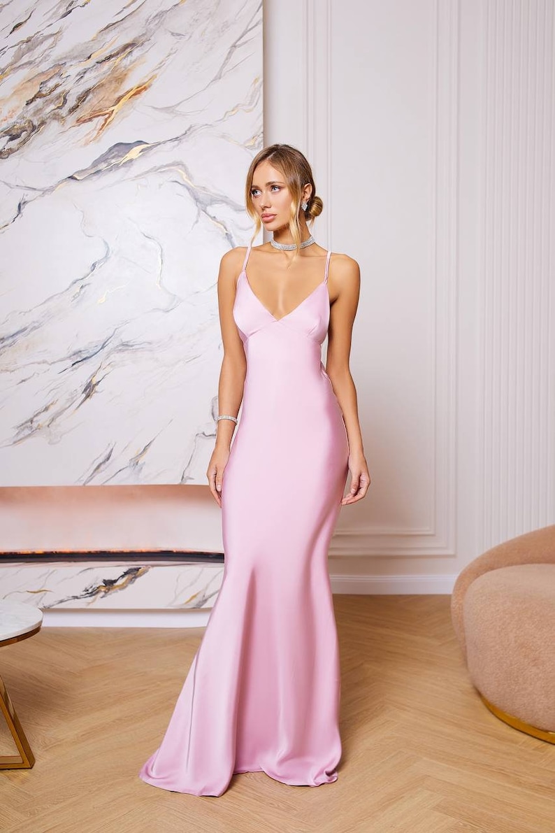 May include: A woman wearing a light pink satin maxi dress with a V-neckline and spaghetti straps. The dress has a fitted bodice and a flowing skirt.
