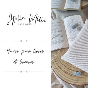 Op de afbeelding: Een beige boekomslag met de tekst "Atelier Millie Hand Made" en "Housse pour livres et liseuses" in zwart. De omslag is gemaakt van geribbeld materiaal en heeft een subtiele textuur.