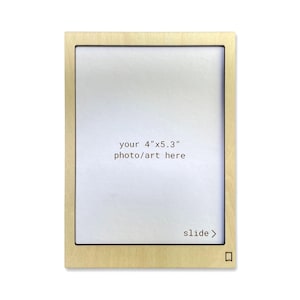 Może przedstawiać: Jasna drewniana ramka na zdjęcia z otworem przesuwnym na zdjęcie lub dzieło sztuki o wymiarach 10,2 cm x 13,5 cm. Ramka ma strzałkę "przesuń" i mały kwadratowy wycięcie w prawym dolnym rogu.
