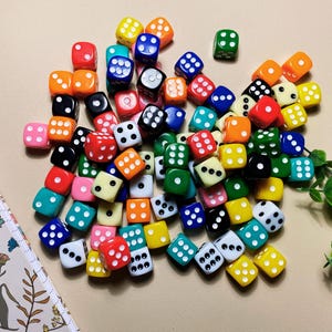 May include: A collection of colorful dice in various colors, including red, green, blue, yellow, and orange, with white pips. The dice are scattered on a beige surface, next to a floral patterned notebook and green leaves.