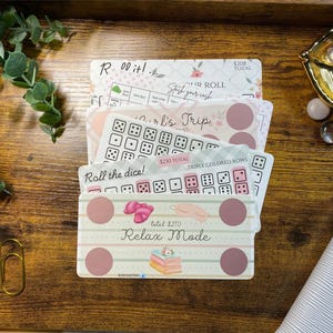 May include: A stack of five rectangular paper savings challenge trackers with various designs. The top card features the text Relax Mode, illustrations of a sleep mask, slippers, and books, with four circular spots and a total of $270 printed on it.
