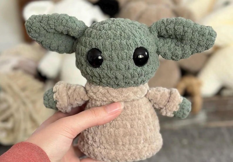 May include: A handmade crochet doll in the likeness of a popular character. The doll has large black eyes, large green ears, and is wearing a tan-colored outfit. The doll is approximately 6 inches tall.