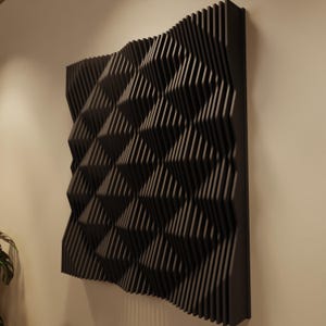 May include: A dark gray, geometric wall panel with a repeating pattern of angled, vertical lines. The panel is mounted on a light-colored wall, creating a contrast. The design has a 3D effect, adding depth and texture to the wall decor.