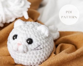 May include: White crocheted cat amigurumi toy with black eyes and a pink nose. The toy is sitting on a brown fabric surface. PDF PATTERN.