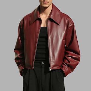 Può includere: Giacca in pelle bordeaux con ampio colletto e cerniera argentata. La giacca ha maniche lunghe e una vestibilità corta. La persona nell'immagine indossa una canotta nera e pantaloni neri.