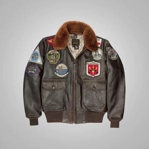 Puede incluir: Una chaqueta bomber de cuero marrón con cuello de piel de oveja y varios parches. Los parches incluyen un emblema de la Fuerza Aérea de los Estados Unidos, un parche de escuadrón y una etiqueta con el nombre.