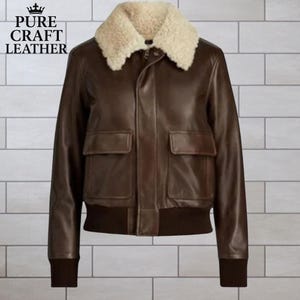 Könnte beinhalten: Braune Leder-Bomberjacke mit cremefarbenem Shearling-Kragen. Die Jacke hat zwei Vordertaschen und einen gerippten Saum und Bündchen. Der Text "PURE CRAFT LEATHER" befindet sich oben links.