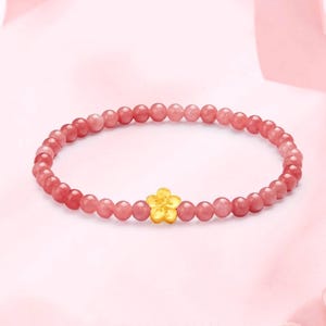 May include: A bracelet made of small, round, pink gemstones, with a gold-coloured flower charm at its centre. The bracelet is set against a soft pink backdrop.