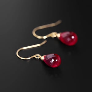 Puede incluir: Un par de pendientes colgantes dorados con gemas rojas facetadas en forma de lágrima. Los pendientes se muestran sobre un fondo oscuro, resaltando su color y diseño. Los ganchos son curvos y sencillos.
