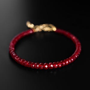 May include: A red beaded bracelet with a gold clasp. The bracelet is made of small, faceted red beads. The clasp is a bright gold color. The bracelet is set against a black background.