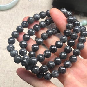 Op de afbeelding: Een verzameling donkergrijze kralenarmbanden. Elke armband is gemaakt van gladde, ronde kralen. De armbanden worden in een hand gehouden, waarbij hun kleur en textuur worden getoond. De kralen hebben een subtiel, gespikkeld patroon.