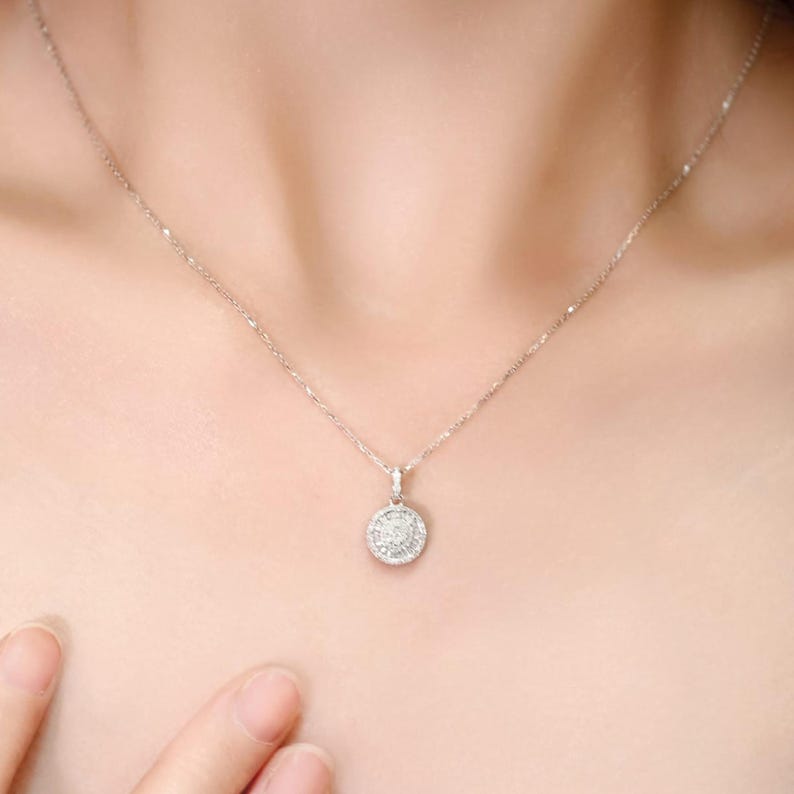 May include: A delicate silver necklace with a round pendant. The pendant is encrusted with small, sparkling stones, creating a halo effect. The necklace is worn around the neck.
