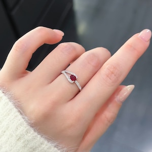 Könnte beinhalten: Ein silberner Ring mit einem roten Edelstein und kleinen Diamanten, die in einem zarten, geschwungenen Design gefasst sind.