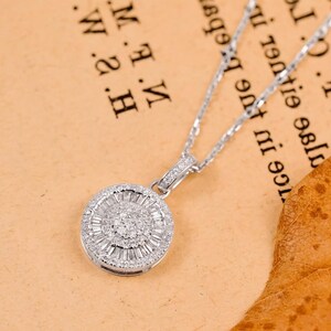 May include: A silver necklace with a round pendant. The pendant is encrusted with small, sparkling stones arranged in a circular pattern. The necklace chain is delicate and also silver. The background is a light brown color.