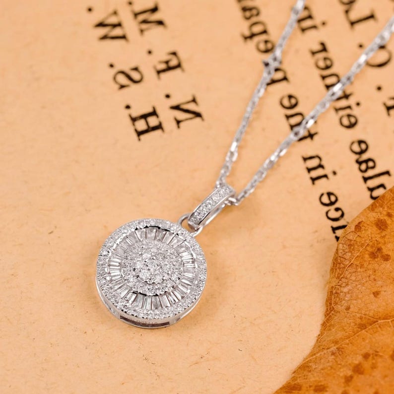 May include: A silver necklace with a round pendant. The pendant is encrusted with small, sparkling stones arranged in a circular pattern. The necklace chain is delicate and also silver. The background is a light brown color.