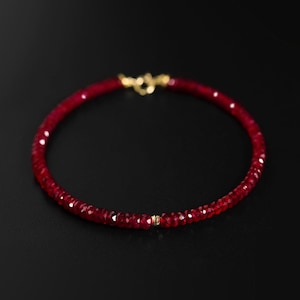 Puede incluir: Una pulsera de piedras preciosas rojas con un cierre dorado. La pulsera está compuesta por pequeñas cuentas rojas facetadas, ensartadas para formar un círculo. Un pequeño acento dorado es visible cerca del centro de la pulsera.
