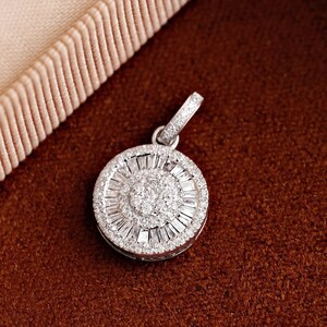 May include: A round, silver pendant encrusted with numerous small, sparkling diamonds. The pendant features a central cluster of diamonds surrounded by a ring of baguette-cut stones. The bail is also adorned with diamonds. The jewelry is set against a brown background.