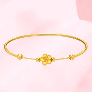 Può includere: Un bracciale rigido dorato con un elemento floreale centrale. Il bracciale presenta un design delicato con un fiore e piccoli accenti a sfera. Lo sfondo è rosa tenue.