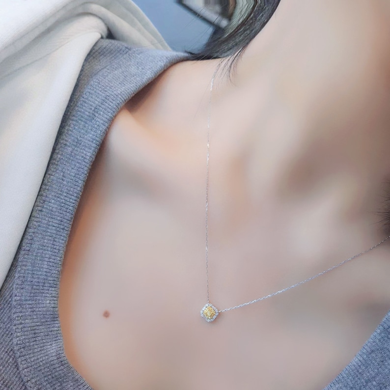 May include: A delicate silver chain necklace with a small square pendant featuring a yellow gemstone surrounded by smaller clear gemstones.