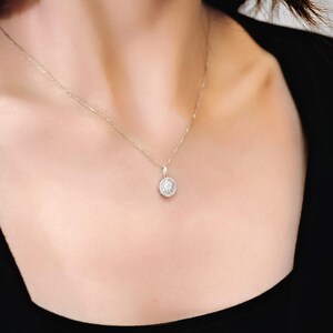 May include: A delicate silver necklace with a round pendant. The pendant is encrusted with small, sparkling stones, creating a halo effect. The necklace is worn against a dark background, highlighting the pendant's brilliance. A classic piece of jewelry.