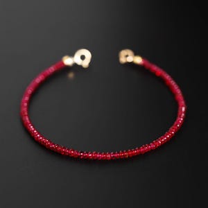 May include: A fine red gemstone bracelet with gold-coloured clasps. The bracelet features small, faceted red beads strung together. The background is a solid black, accentuating the vibrant colour of the bracelet.