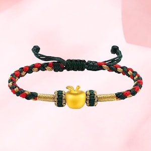 May include: Gold-colored apple charm bracelet with a braided cord in red, green, and gold. The bracelet features a gold apple charm and gold accents. The adjustable cord is green and red.