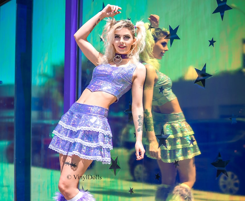 May include: A woman wearing a purple iridescent crop top and matching mini skirt with white lace trim. She is posing against a mirrored wall with black stars. The text "VinylDolls" is visible in the bottom right corner of the image.