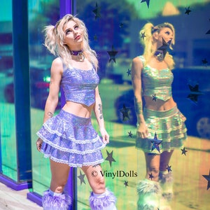 May include: A woman wearing a holographic purple crop top and a layered iridescent skirt with white lace trim. She is wearing white furry boots and has her hair in pigtails. The background is a mirrored wall with stars.