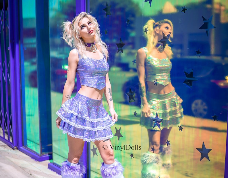 May include: A woman wearing a holographic purple crop top and a layered iridescent skirt with white lace trim. She is wearing white furry boots and has her hair in pigtails. The background is a mirrored wall with stars.