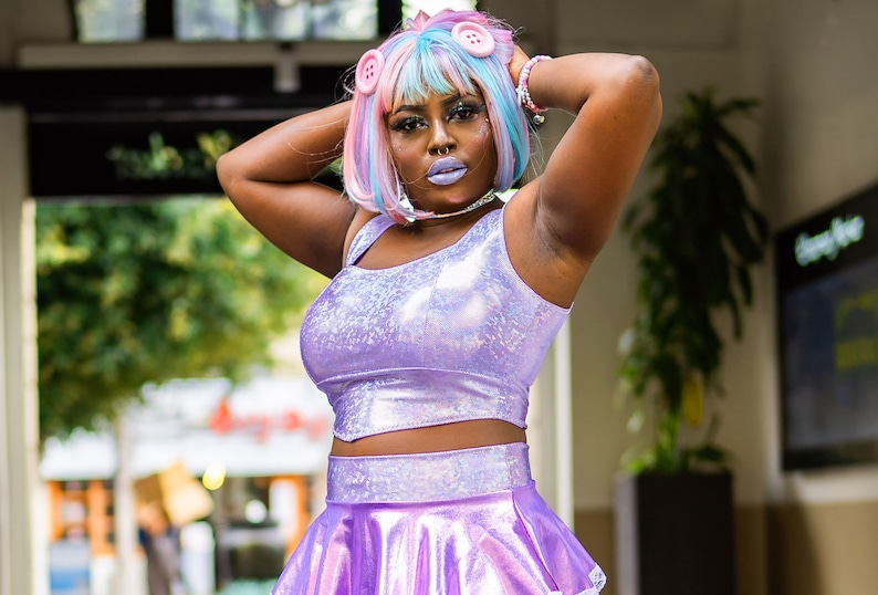 May include: A woman wearing a purple iridescent crop top and skirt with a pink and blue wig with buttons.