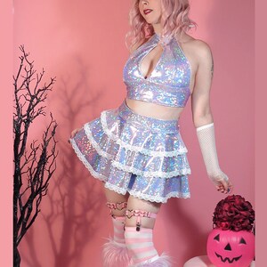 May include: A woman wearing a silver iridescent halter top and a layered skirt with white lace trim. She is standing in front of a pink background with a black tree branch in the background. She is holding a red rose and a jack-o-lantern.