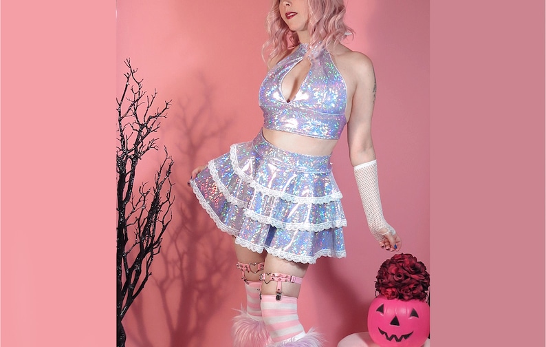 May include: A woman wearing a silver iridescent halter top and a layered skirt with white lace trim. She is standing in front of a pink background with a black tree branch in the background. She is holding a red rose and a jack-o-lantern.