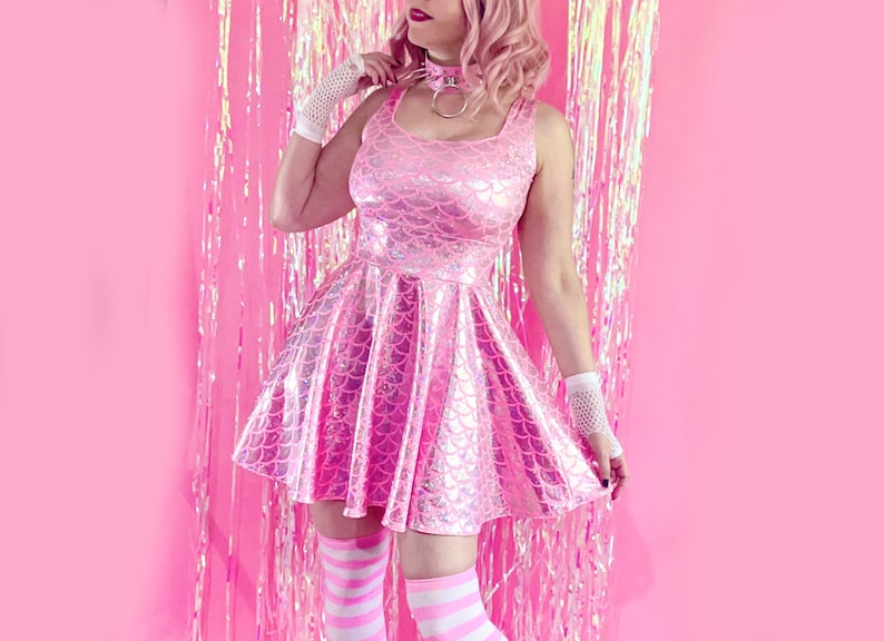 May include: A pink iridescent dress with a mermaid scale pattern. The dress has a fitted bodice and a flared skirt. The model is wearing white fishnet gloves and white and pink striped socks.