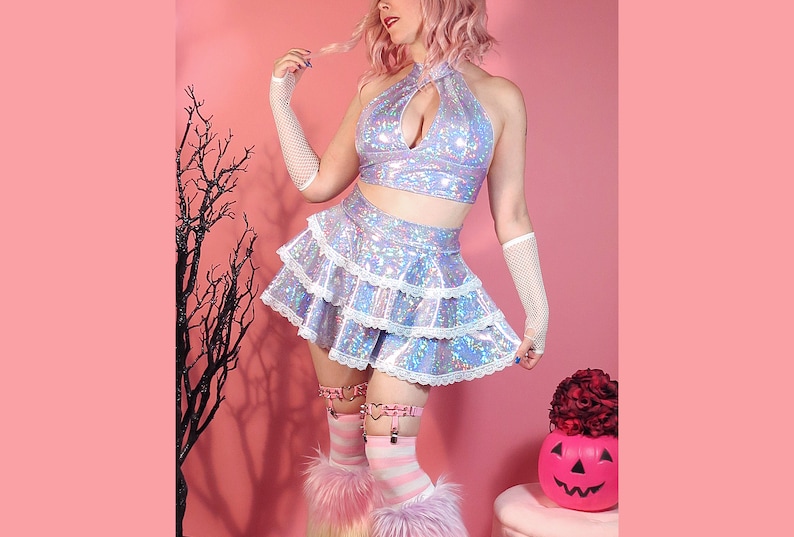May include: A holographic iridescent crop top and skirt set with a white lace trim. The top has a halter neckline and a keyhole cutout in the front. The skirt is a mini skirt with multiple layers of ruffles.