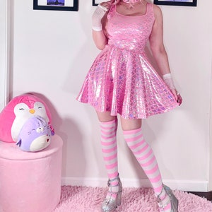 May include: A pink iridescent dress with a mermaid scale pattern. The dress has a scoop neck and is sleeveless. The model is wearing white striped knee-high socks and silver platform shoes.